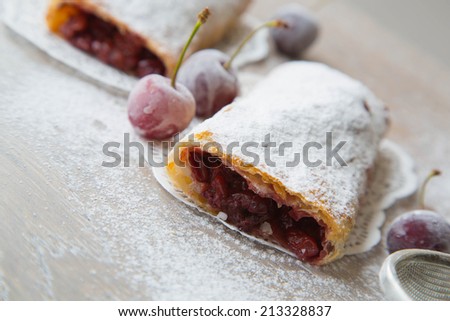 Traditional romanian dessert - invertita.Baked rolled thin dough stuffed with sour cherries. Frozen sour cherries and a tea strainer in the background.The dessert placed on the wooden surface.Close up