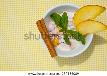 Homemade peach yogurt in a white dish on a yellow table cloth. Decorated with two slices of fresh peach and fresh peppermint leaves and cinnamon stick. Top view.