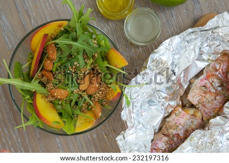 Vitamin salad- rucola with almond, peach and sesame seeds in glass dish and baked fish in the aluminium foil from the right side. Top view