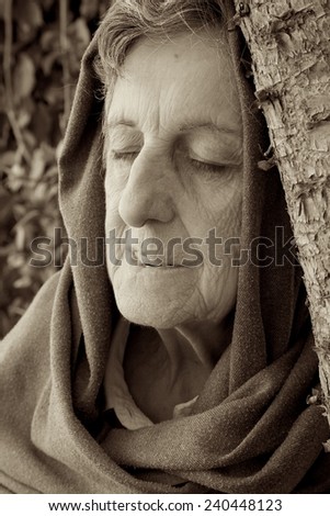 An old woman between 70 and 80 years old is recollecting happy moments of her life with closed eyes having leant against a trunk of the old tree