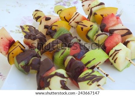 Fitness dessert: fruit salad skewers covered in bitter chocolate. Fruits in the background.