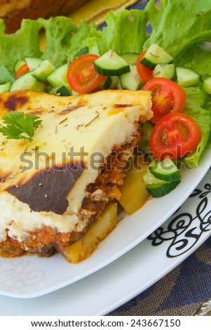 Traditional Greek dish - moussaka - aubergines and potatoes based dish. Served with fresh salad leaves,cherry tomatoes and cucumbers. Glass mold with entire dish is in the background.