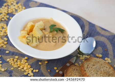 Vegetarian light lunch: cream soup with yellow lentils, fresh parsley and croutons in a white round deep plate. Spoon with wooden handle in the background