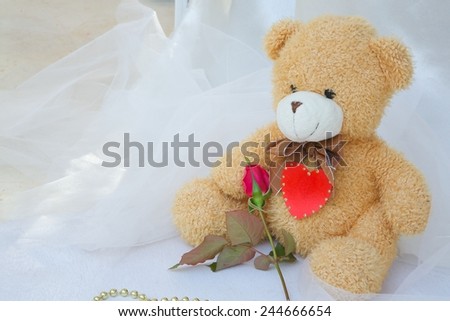 Brown plush Teddy bear with red heart and red rose. Golden like perls on a white wooden surface.