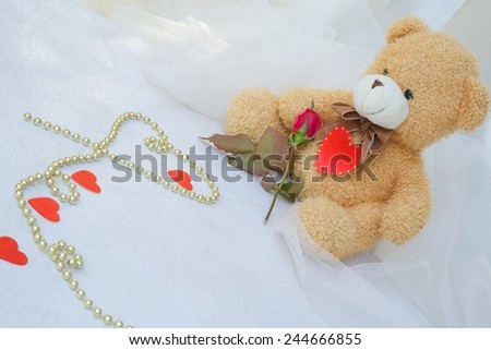Brown plush Teddy bear with red heart and red rose. Golden like perls on a white wooden surface.