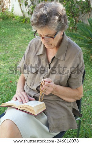 A senior woman between 70 and 80 years old is praying in the garden, having Franciscan prayer book on her knees and Christian prayer beads with cross on her knees