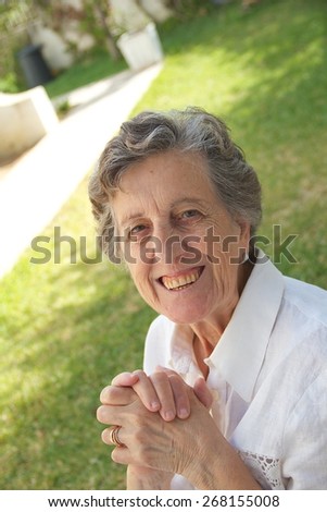 A happy smiling old woman between 70 and 80 years old is keeping two hands together sitting in the garden