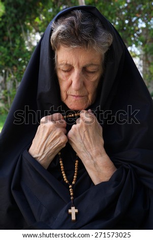 A senior woman between 70 and 80 years old, dressed in black is praying in the garden. Fron view