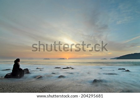 An unidentified person sitting on a boulder watchig sunset while waves hit the beach.