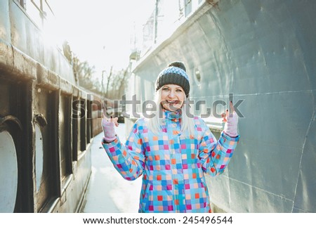 Smiling naughty blonde woman in bright jacket and hat with pompom doing rock and roll gesture in sunny winter outdoors