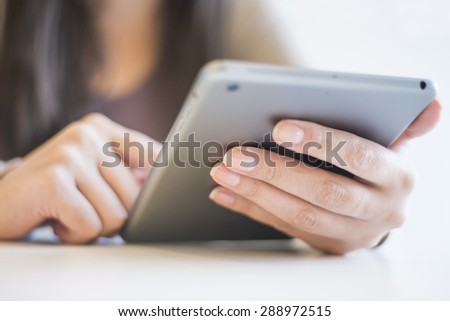 Woman is holding a digital tablet. The image is taken in the city. The young woman has long brown hair. The face of the  woman is not visible.