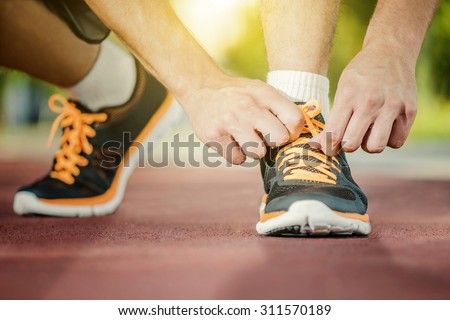 A person running outdoors on a sunny day. Only the feet are visible. The person is wearing black running shoes.Focus on a side view of two human hands reaching down to a athletic shoe.