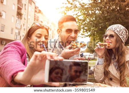 Group of young people laughing and doing a selfie in cafe.They are eating pizza and having a great time. It is a nice day with sunshine.Selective focus