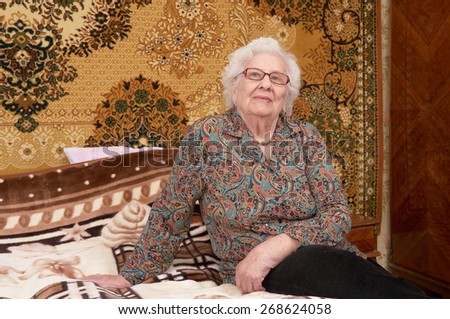 Portrait of happy senior caucasian woman in glasses about ninety years old sitting on bed in her bed room