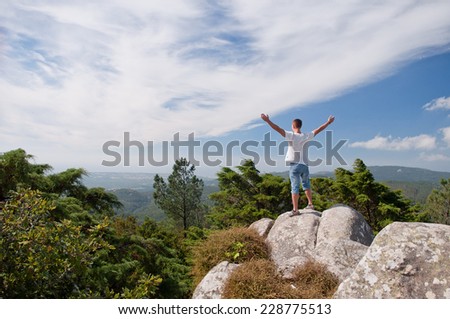 Young happy man standing on a rocks with raised hands and looking to the landscape below in Portugal
