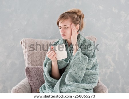 Attractive woman with natural blond hair posing without makeup with headache and white cup of hot drink wrapped with blanket at home