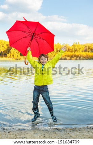 Happy little boy in rubber boots and raincoat hopping and making splashes in water