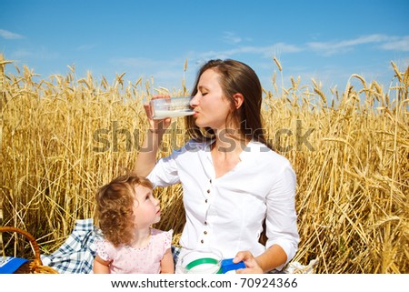 Woman drinking milk in wheat field, curly daughter looking at her