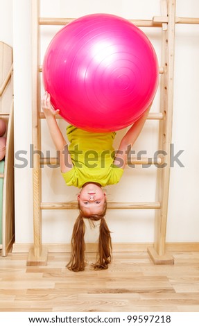 Elementary aged girl hanging on wooden wall bars with a large gymnastic ball