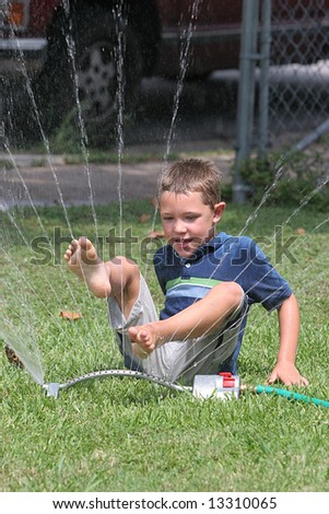 stock photo Young boy playing in the sprinkler with his feet in the air