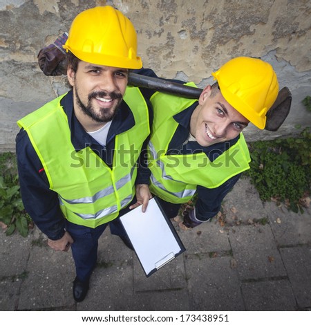 Two manual workers happy with their working conditions smiling, in protective suites, one holding paper and pen, the other one holding big hammer. View from above.