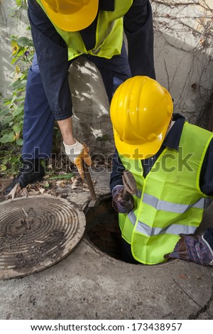 Two young manhole workers, one of them being in the sewer looking for problem to fix.