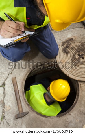 Two manual hard workers on field writing down the water inside of manhole at the road. Detail, view from above.