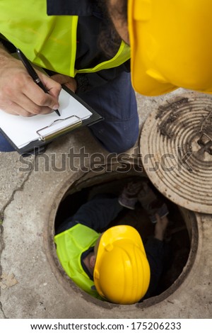 Two manual hard workers on field writing down the water inside of manhole at the road. Detail, view from above.
