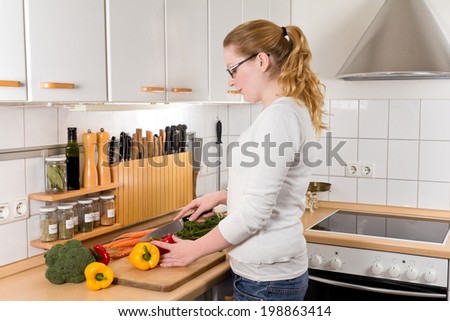 Slim young woman with glasses cutting vegetables in her kitchen, side view