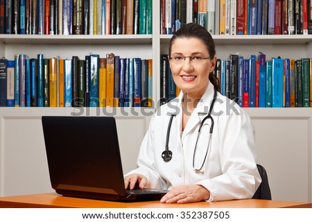 Middle aged or mature female doctor sitting at her desk in front of a computer and smiling friendly