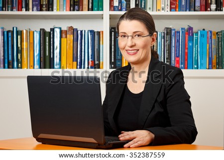 Friendly smiling woman in front of her laptop in a library, teacher, tutor, professor, consultant, consultancy