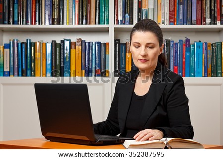Mature woman in an office or library with her laptop reading in a book, advocate, lawyer, teacher, prof, historian, psychologist, diplomat