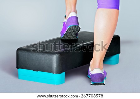 Female feet in violet sneakers do exercise on a black-turquoise fitness aerobic step. Rear view.