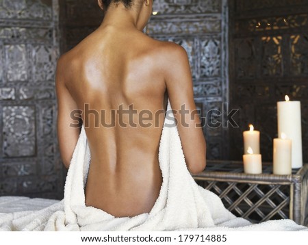 Naked back of a young Asian woman meditating sitting on a bed in front of lit candles on a small cabinet with a towel draped over her front
