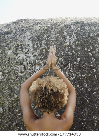 View from behind of the bare head and shoulders of a young Asian woman practising yoga meditating with her hands raised above her head against a granite rock