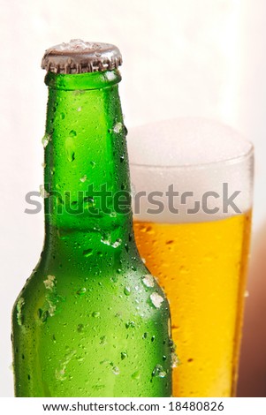 Green bottle of beer in front with drops and chips of ice with a glass of beer in the back with drops and foam