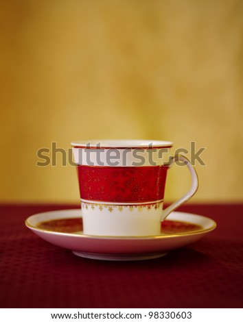 Vintage coffee cup with red and gold patterns over red carpet against yellow background a small coffee jar is blurred in the background. Dynamic angle accentuate the cup and give motion to the scene.