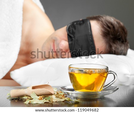 Man with Sleeping mask sleep on a bed, cup of herbal tea in the foreground. Focus on tea cup
