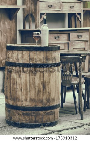 Vintage stylized photo of wooden barrel with bottles of wine and glass, chair and table in outdoor cafe