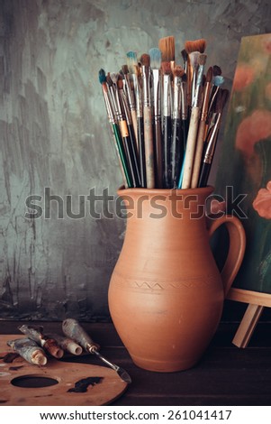 Paintbrushes in a jug from potters clay, palette, paint tubes and Painting in artist studio.