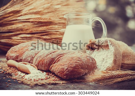 Fresh bread, jug of milk, sack with flour and sheaf of wheat on wooden table. Retro stylized.