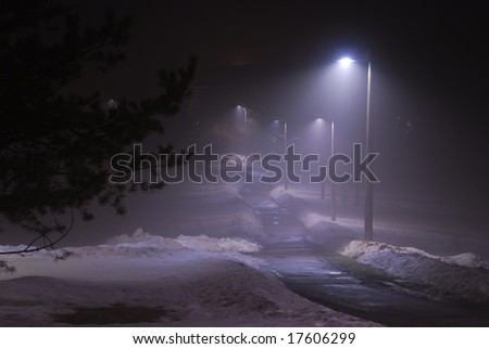 A light fog is lit up by the lights along a snowy walk at night.