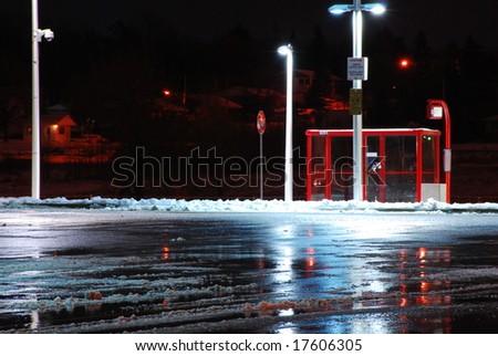 A bus stop on a cold winter night sits empty.  Lights reflect off the wet parking lot pavement.