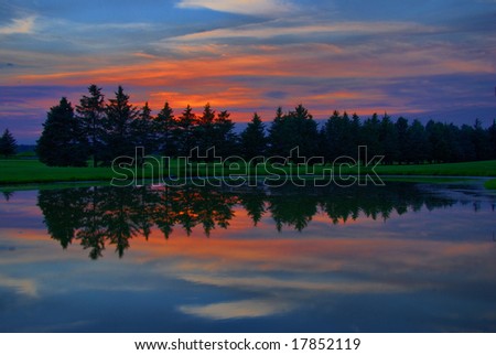 The sky and trees reflect off the water at sunset on a golf course.