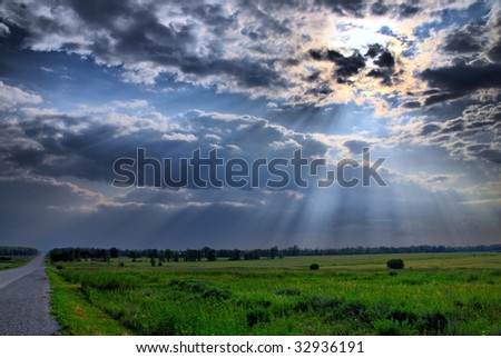 Sun beams break through the clouds over farmland by a country road.