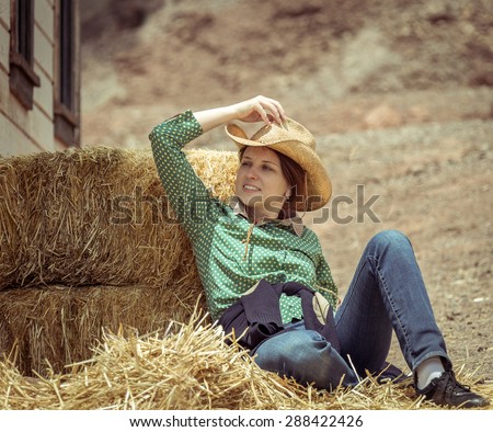 Cowgirl woman smiling happy sitting next to haystack wearing cowboy straw hat. Beautiful young Caucasian girl in countryside