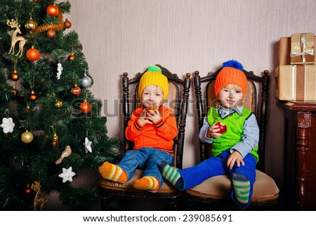 Little brother and sister sitting on chairs near Christmas tree and eat apples