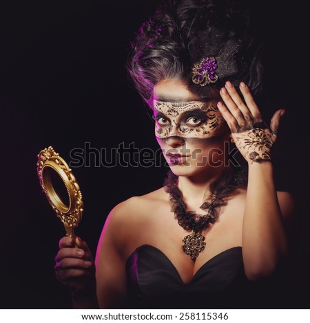 A mysterious young woman in carnival mask with an unusual makeup looks in the mirror