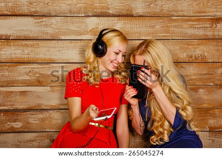 Two sisters, blonde, listening to music on headphones. Girls having fun, holding a cell phone and a tablet pc. Sisters sitting in the bedroom.