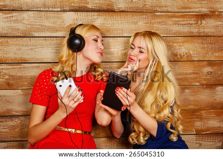 Two sisters, blonde, listening to music on headphones. One girl sends an air kiss another. Girls are holding a smartphone and a tablet pc. Sisters sitting in the bedroom.